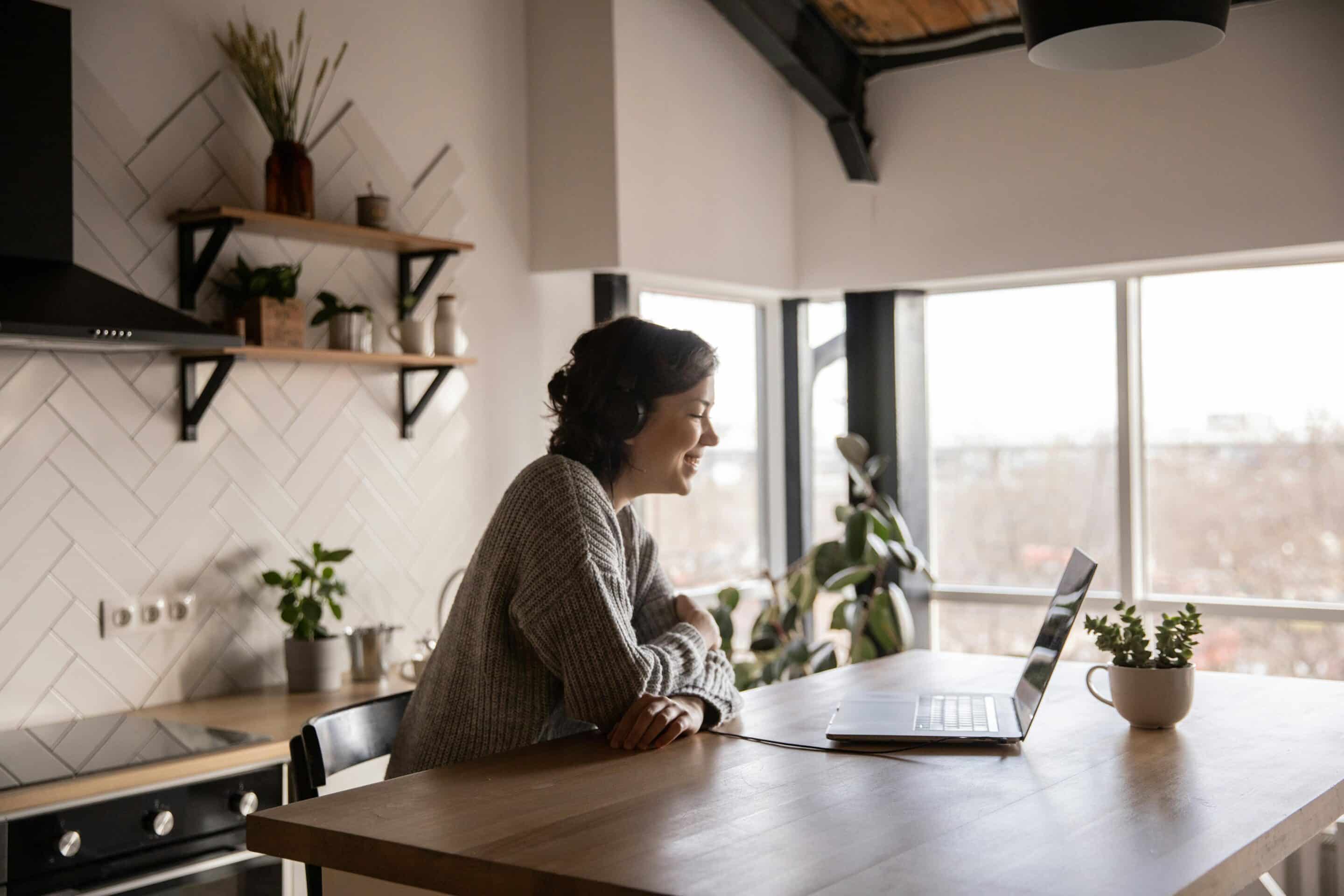 A person wearing headphones sits at a wooden kitchen island, smiling while looking at a laptop during a video call. The bright, modern kitchen features large windows, white tile walls, open shelving, and several potted plants.