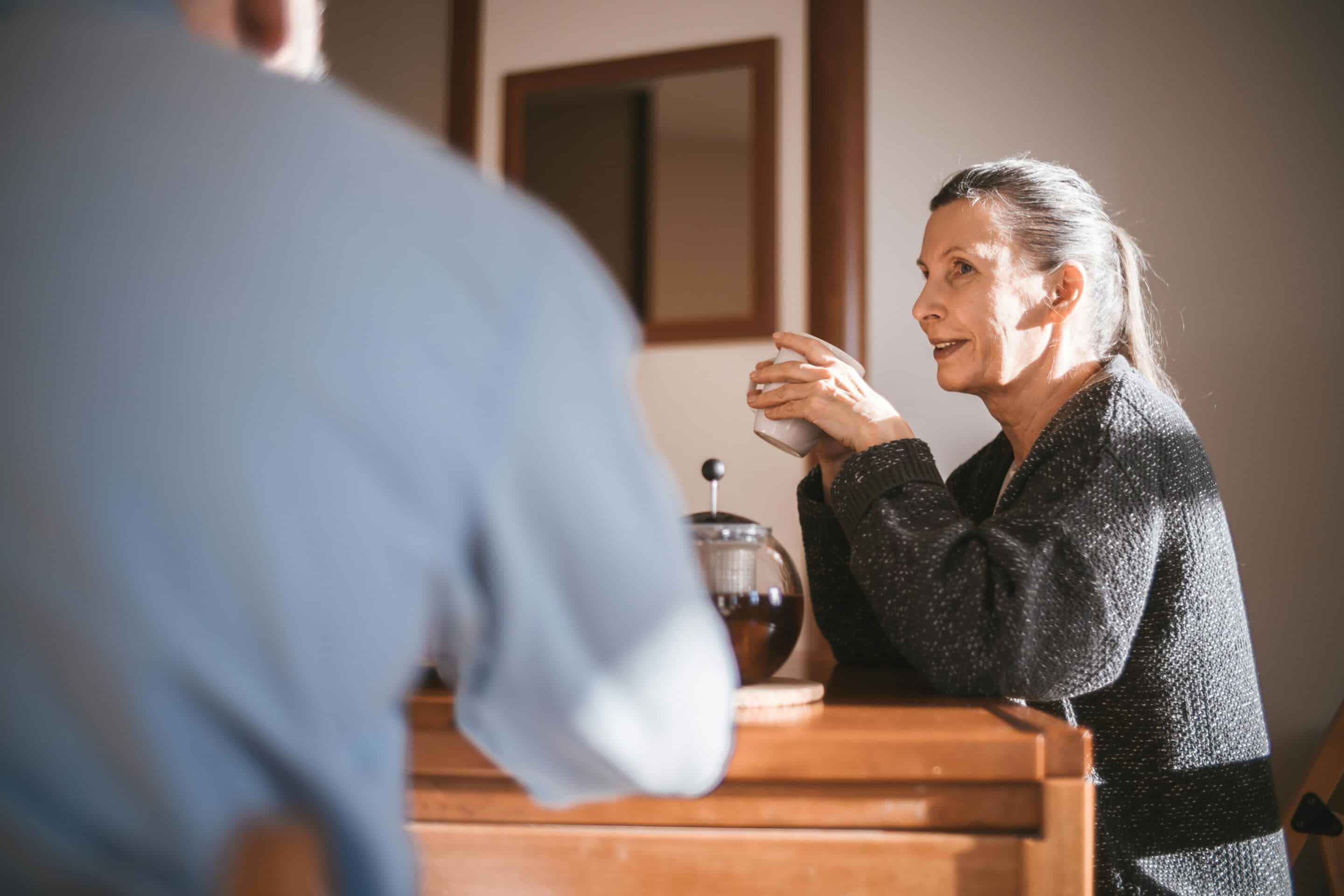 A resident in a dark cardigan holds a coffee mug while having a friendly conversation with a property team member in a light blue sweater, in a warm apartment kitchen setting with a French press and wooden counter — representing personal, community-focused resident engagement.